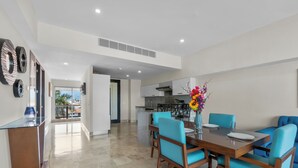 A modern dining area with a wooden table and blue chairs, a kitchen in the background, and a view of the outdoors. - 3rd-Floor Gem w/ Cruise Views & Rooftop Lounge (Puerto Vallarta)