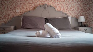 Ein ordentlich bezogenes Bett mit zwei zusammengerollten Handtüchern, einem Kopfteil, zwei Nachttischen mit Lampen und floralem Wandpapier.