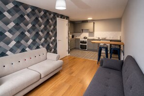 A compact living space with a sofa, a dining table with stools, a kitchen area, and a patterned wall.