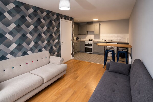 A compact living space with a sofa, a dining table with stools, a kitchen area, and a patterned wall.