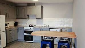 A compact kitchen with grey cabinets, a white stove, a microwave, a sink, and a wooden table with stools. - Central Apartment in Belfast City Centre (Belfast)