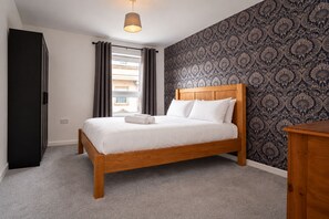 A bedroom with a wooden bed, grey curtains, a patterned wallpaper, and a small wooden nightstand.
