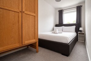 A bedroom with a wooden wardrobe, a bed with white bedding, a nightstand with a lamp, and a window with curtains.