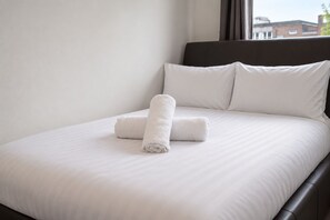 Una cama bien tendida con sábanas blancas y dos toallas enrolladas, un cabecero oscuro y una ventana con vistas a edificios.