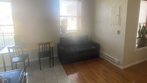 A living room with a dark-colored sofa, a wooden dining table with chairs, a potted plant, and a window with blinds. - Pet Friendly Apartment In Arena District with Parking (Detroit)