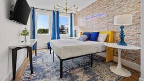 A modern hotel room with a large bed, a TV mounted on the wall, a desk, and a blue and white patterned rug.