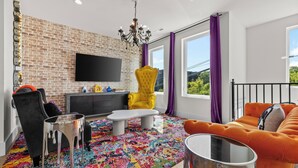 A modern living room with a brick wall, a flat-screen TV, a yellow armchair, a black sofa, and a colorful rug.