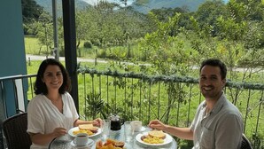 Dos personas sentadas en una mesa redonda de vidrio, con platos de comida y bebidas, con vista a un paisaje exuberante y una montaña al fondo.