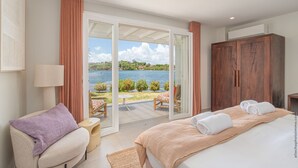 Un dormitorio con una cama grande, una zona de estar con una silla, un armario de madera y vistas a un lago a través de una puerta de cristal.