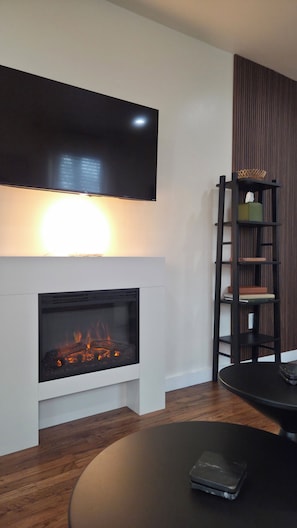 A modern living room with a flat-screen TV, a fireplace with visible fire, a round table, and a bookshelf.