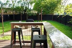A backyard with a wooden table, two black stools, a grill, and a string of lights.