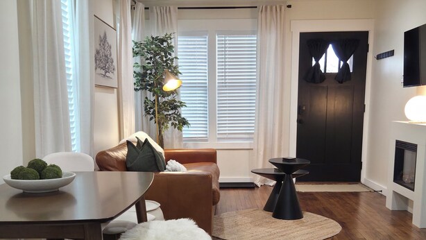 A modern living room with a wooden floor, a brown leather sofa, a dining table with chairs, a potted plant, and a fireplace.