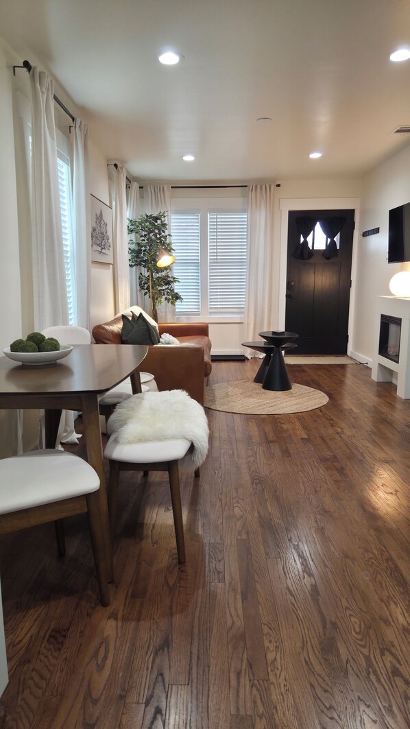 A modern living room with a wooden floor, a brown leather sofa, a dining table with chairs, a potted plant, and a fireplace.