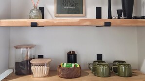 A shelf with a woven basket, a framed sign, a vase, a candle holder, a jar, a wooden bowl with tea bags, and green mugs.