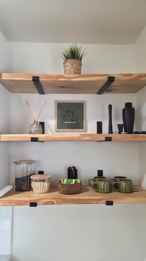A shelf with a woven basket, a framed sign, a vase, a candle holder, a jar, a wooden bowl with tea bags, and green mugs.
