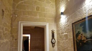 Un couloir aux murs de pierre, avec une porte donnant sur une autre pièce, un luminaire fixé au mur et un tableau encadré.