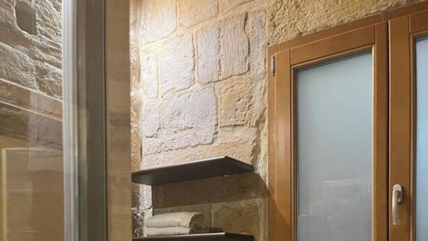 Une salle de bain avec un mur en pierre, des étagères en bois où sont posées des serviettes, un meuble-lavabo en bois avec un vasque, et un miroir avec un cadre en bois.