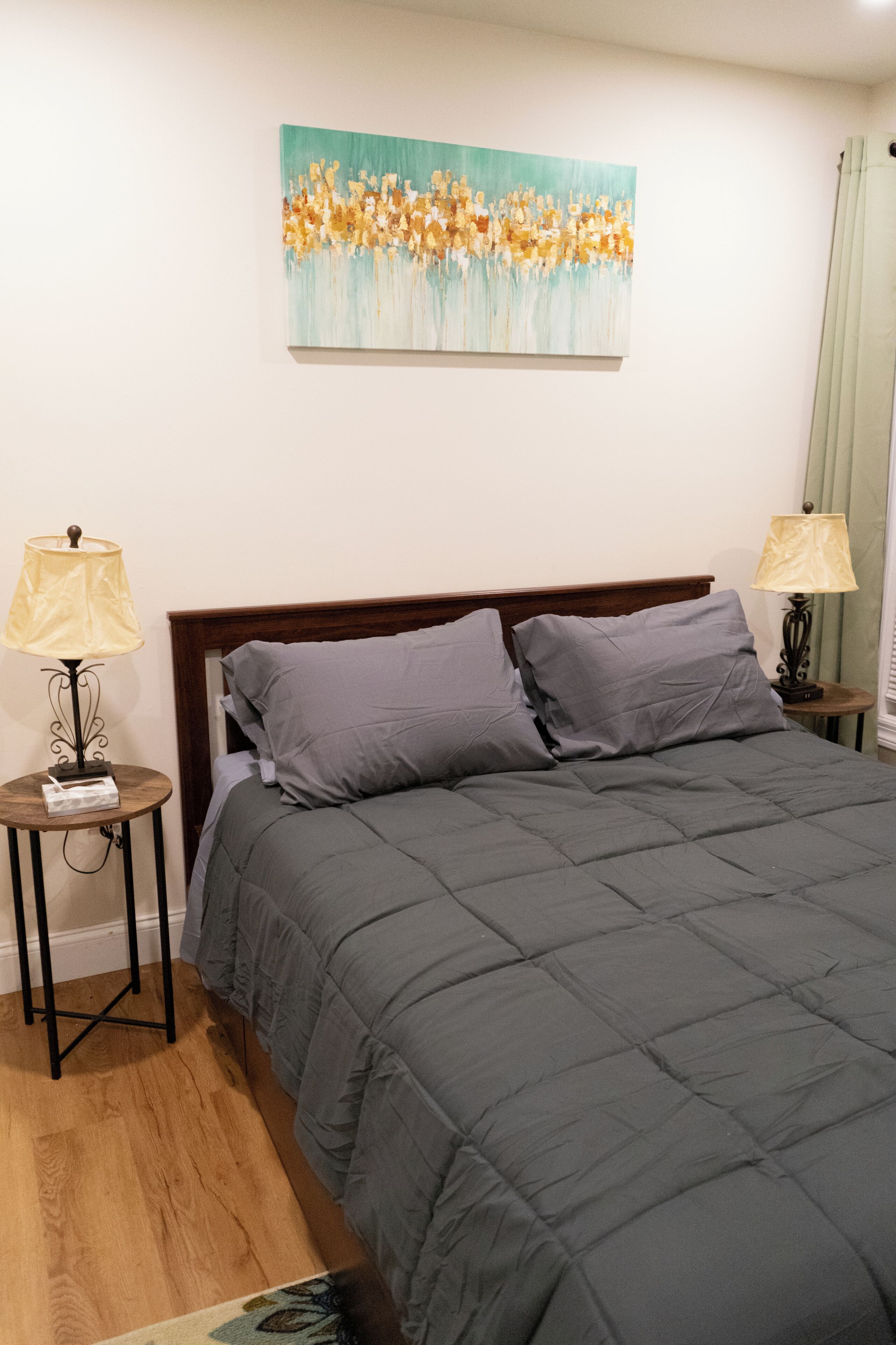 Un lit bien fait, avec une couette grise, deux tables de chevet avec des lampes et un tableau accroché au mur.