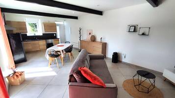A modern living room with a sofa, a dining table with chairs, a kitchen area, and a small black table with a remote control.