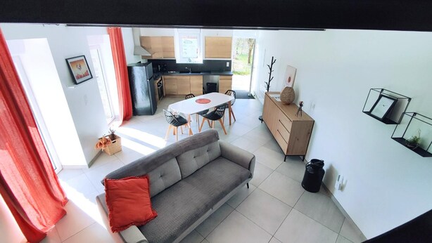 A modern living room with a grey sofa, a dining table with chairs, a kitchen area with a stove and cabinets, and a small side table with decorative items.