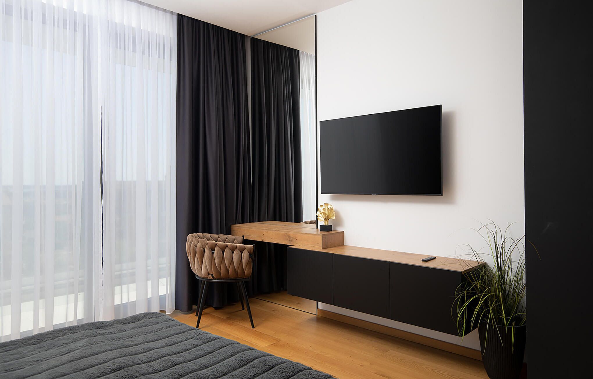 A modern bedroom with a large window, a bed, a bedside table, a chair, a TV, and a plant.
