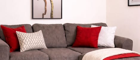 A grey sectional sofa with red and white cushions, a white throw blanket, and a framed abstract tree artwork on the wall.