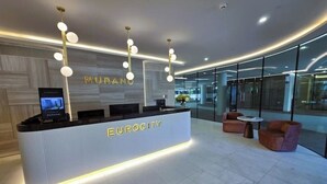 Ein moderner Empfangsbereich mit einem weißen Empfangstresen mit dem Schriftzug "EUROCITY" und einem "MURANO"-Schild darüber. Drei Pendelleuchten befinden sich über dem Tresen, und ein Flachbildfernseher ist an der linken Wand montiert. Der Raum ist gut von Tageslicht durch große Fenster erhellt, und im Vordergrund stehen zwei runde Tische mit Stühlen.