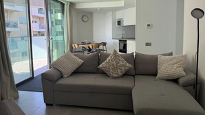 Ein modernes Wohnzimmer mit einer grauen Eckcouch, einem Essbereich mit Holztisch und -stühlen, einer Küche mit weißen Schränken und einer großen Glasschiebetür, die auf einen Balkon führt.