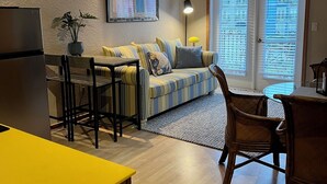 Ein kompakter Wohnbereich mit einer Küchenzeile, einem gestreiften Sofa, einem Esstisch und einem großen Fenster mit Jalousien.