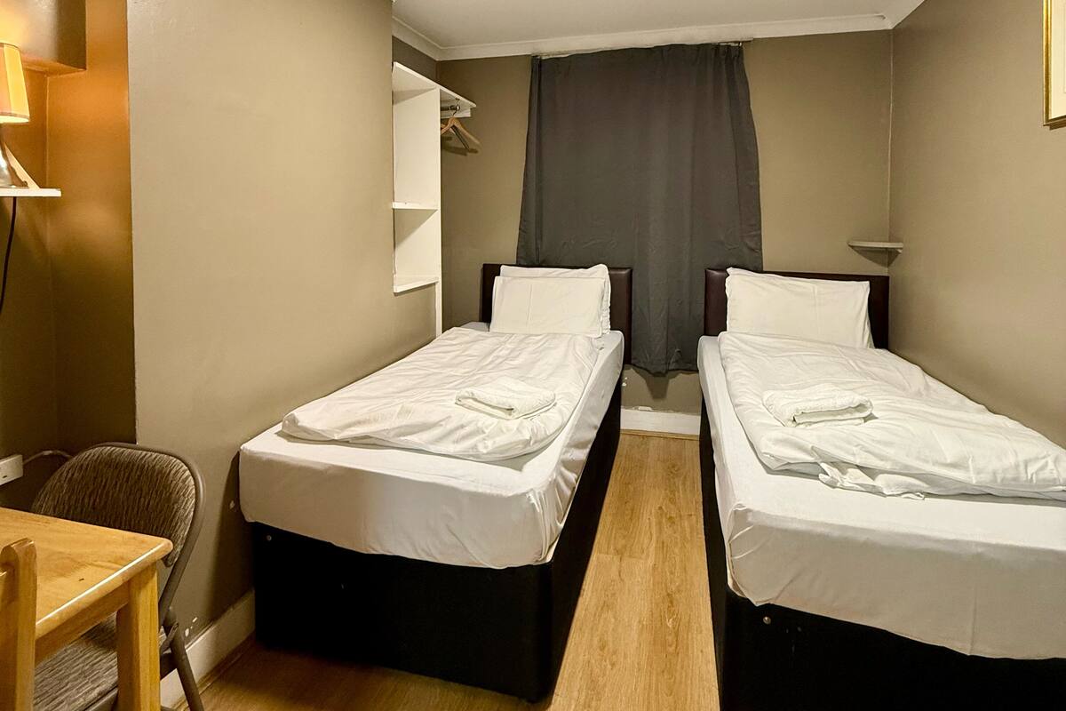 A small, well-lit room with two single beds, a desk, and a chair.