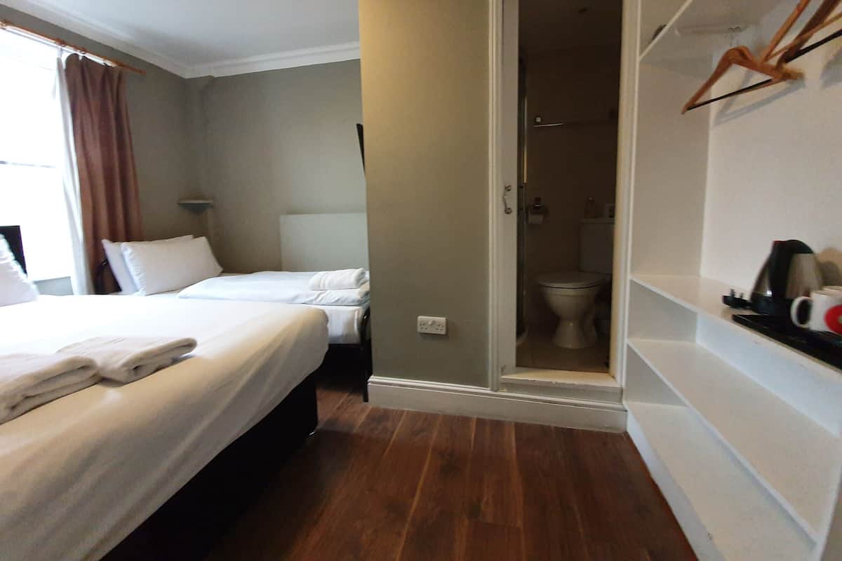 A hotel room with two beds, a shower, a toilet, and a shelf with toiletries.