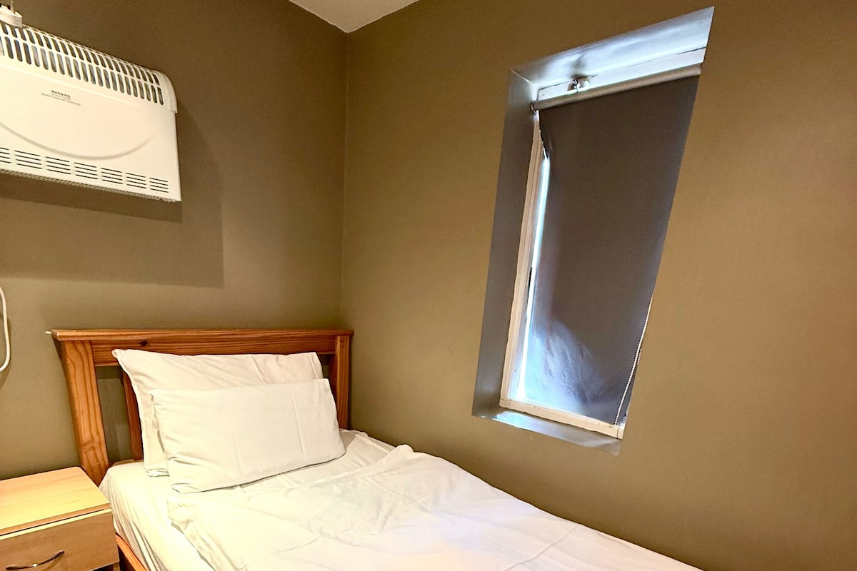 A neatly made bed with a wooden headboard, a bedside table with a lamp, and a window with blinds.