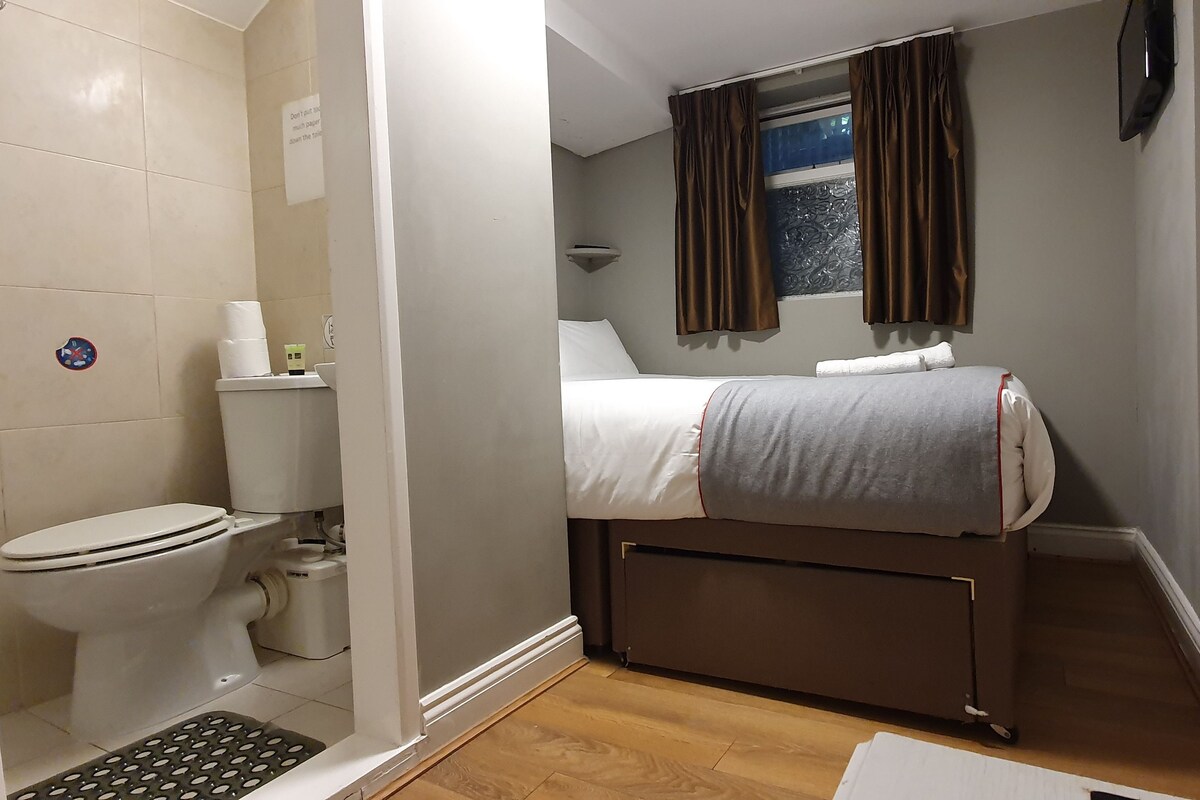 Une chambre d’hôtel de taille réduite, comprenant des toilettes, un petit lavabo, un lit avec une couverture grise, une fenêtre avec des rideaux et un radiateur.