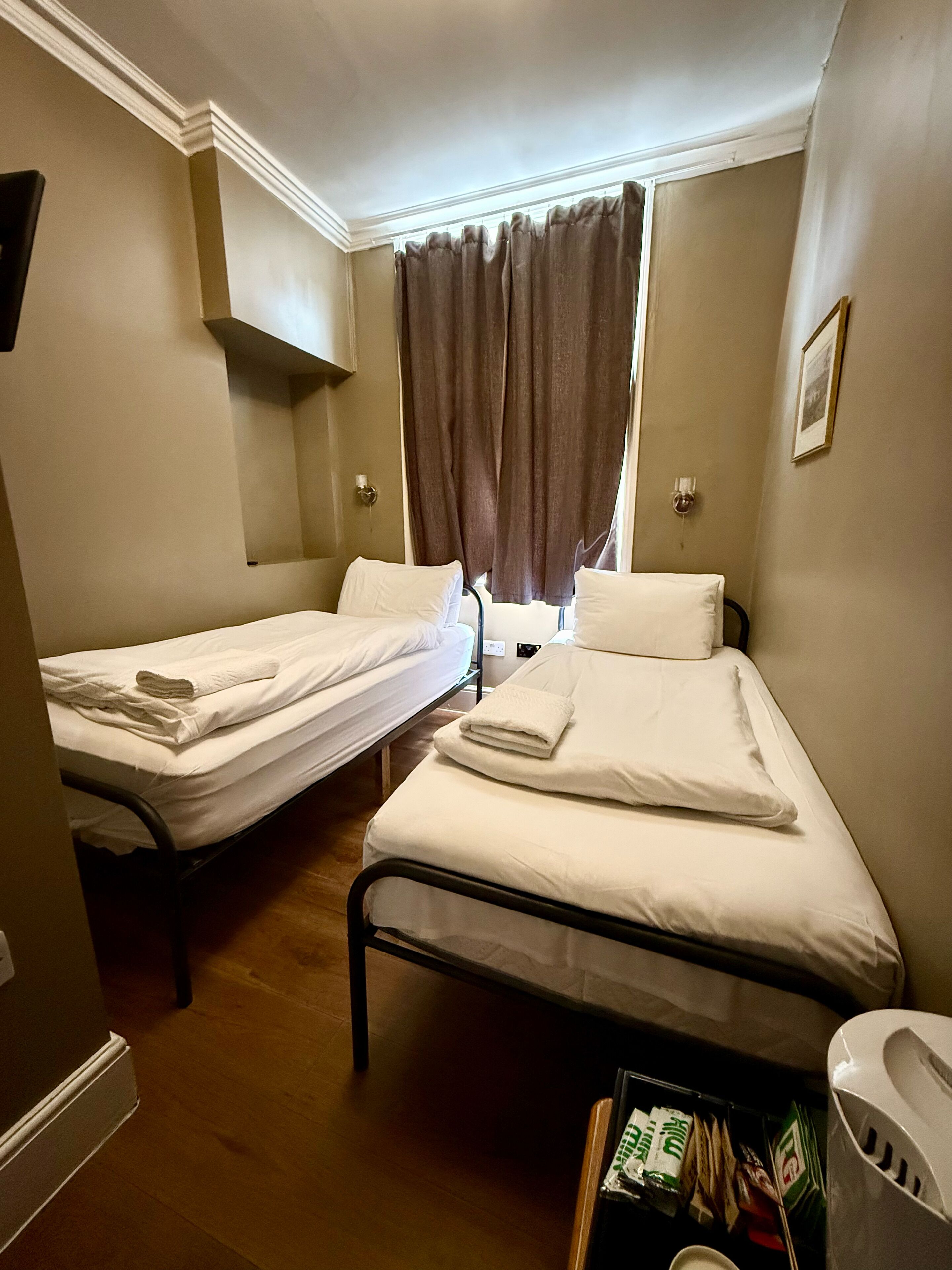 A hotel room with two single beds, a window with curtains, a bedside table, and a small shelf with toiletries.