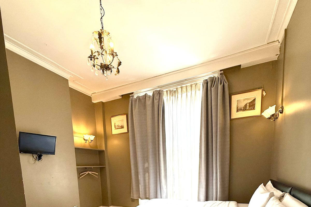A hotel room with a bed, a television, a chandelier, and a window with curtains.