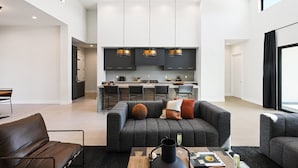A spacious living area with a grey sofa, a coffee table, and a dining table. There is a kitchen in the background with dark cabinets and a bar counter. - Luxe - Solstice Retreat - Heated Pools - 2 Homes (Scottsdale)