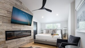 Une chambre à coucher moderne avec une cheminée, un lit, un téléviseur fixé au mur et un ventilateur de plafond.
