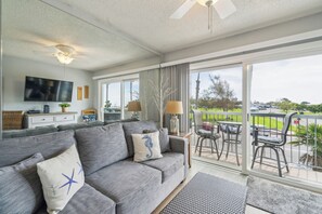 A modern living room with a grey sofa, a flat-screen TV, and a balcony with outdoor seating. - Full Beach Condo with Private Balcony (Carpinteria)