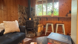 A cozy cabin interior with a stone fireplace, a black sofa, a wooden table with books, and a window overlooking greenery. - Tiny Cabin Couples escape! Hot tub, dry sauna, VIEWS!  (Gold Bar)