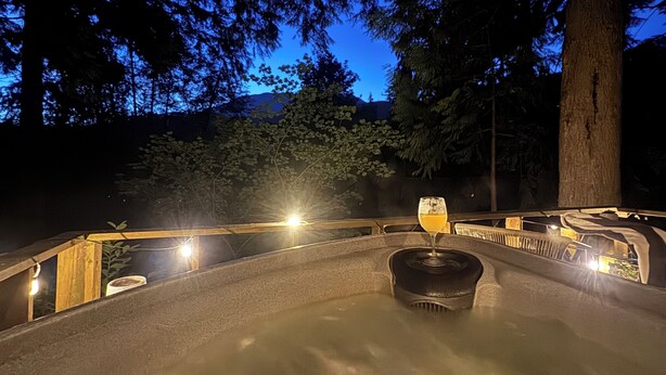 A hot tub area with a glass of orange juice, surrounded by trees and a stone wall, with a view of a mountain in the background. - Tiny Cabin Couples escape! Hot tub, dry sauna, VIEWS!  (Gold Bar)