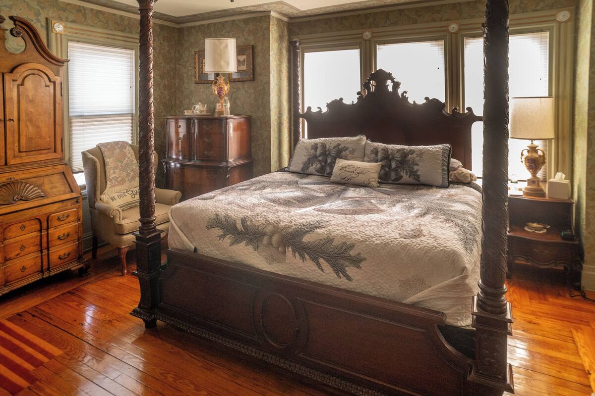 A four-poster bed with a floral bedspread, a wooden dresser, a chair, and a nightstand with a lamp.