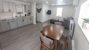 Een moderne keuken met grijze kastjes, een eettafel met stoelen, een grijze bank en een flatscreen televisie aan de muur.