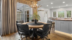 Un coin repas avec une table ronde, des chaises noires, un lustre et une vue sur la cuisine et les fenêtres.