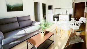 Una sala moderna con un sofá gris, una mesa de centro y un comedor con sillas blancas. Cuenta con una cocina con armarios blancos y un microondas.