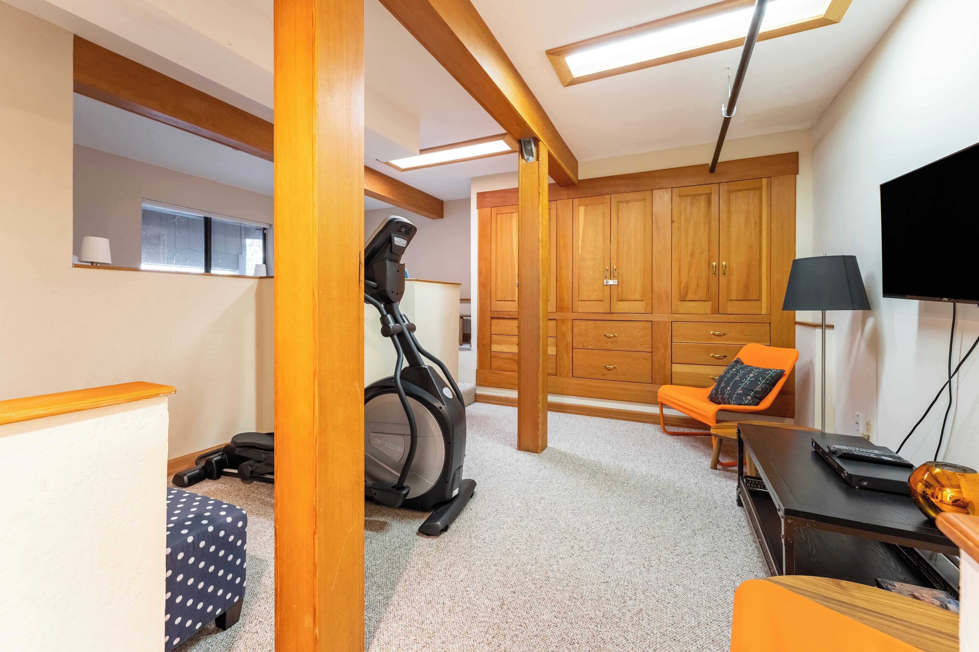 A room with a treadmill, a desk with a laptop, an orange chair, and wooden cabinets.