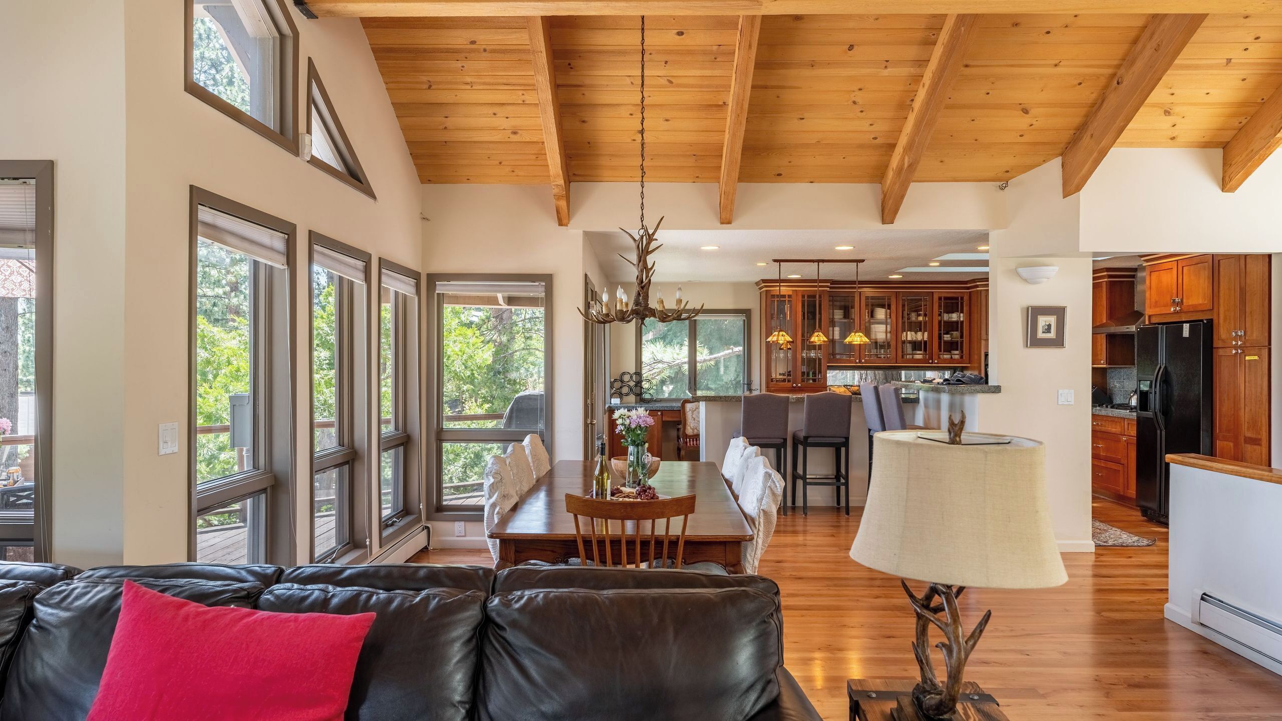 A spacious living room with a black leather couch, a dining area with a table and chairs, a kitchen with wooden cabinets, and a ceiling with exposed beams.