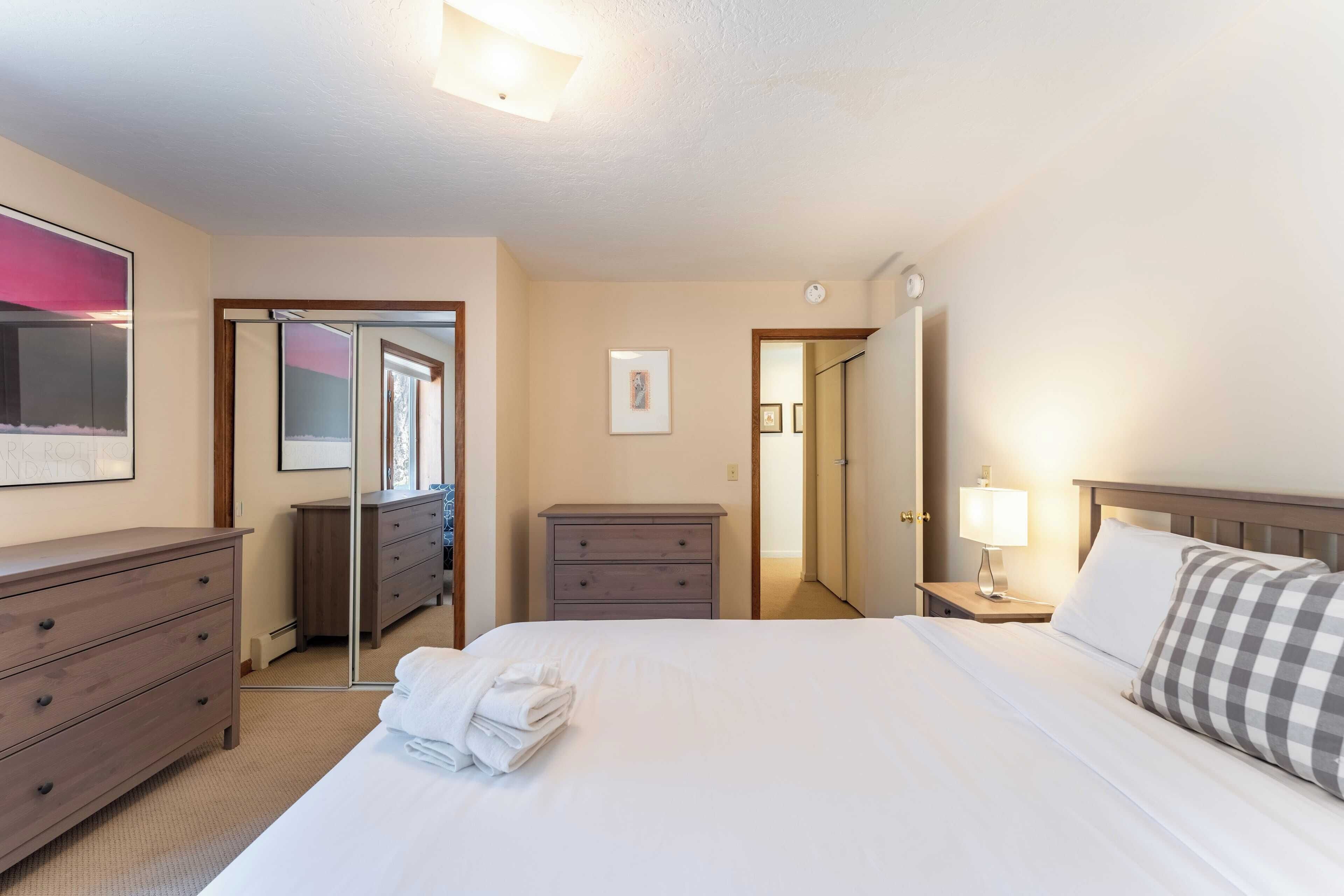 A hotel room with a bed, a dresser, a nightstand with a lamp, a mirror, and a door leading to another room.