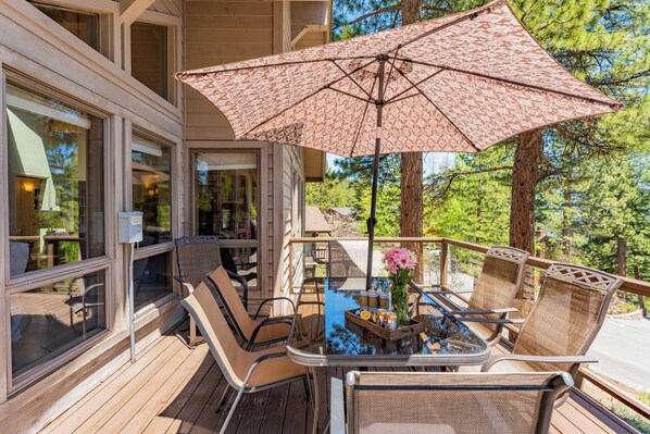 A wooden deck with wicker chairs and a table, a large patterned umbrella, and a view of a forested area.