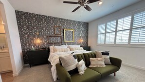 Une chambre à coucher avec un lit, un canapé vert, un ventilateur de plafond, une fenêtre avec des stores et un mur recouvert d’un papier peint à motifs floraux.