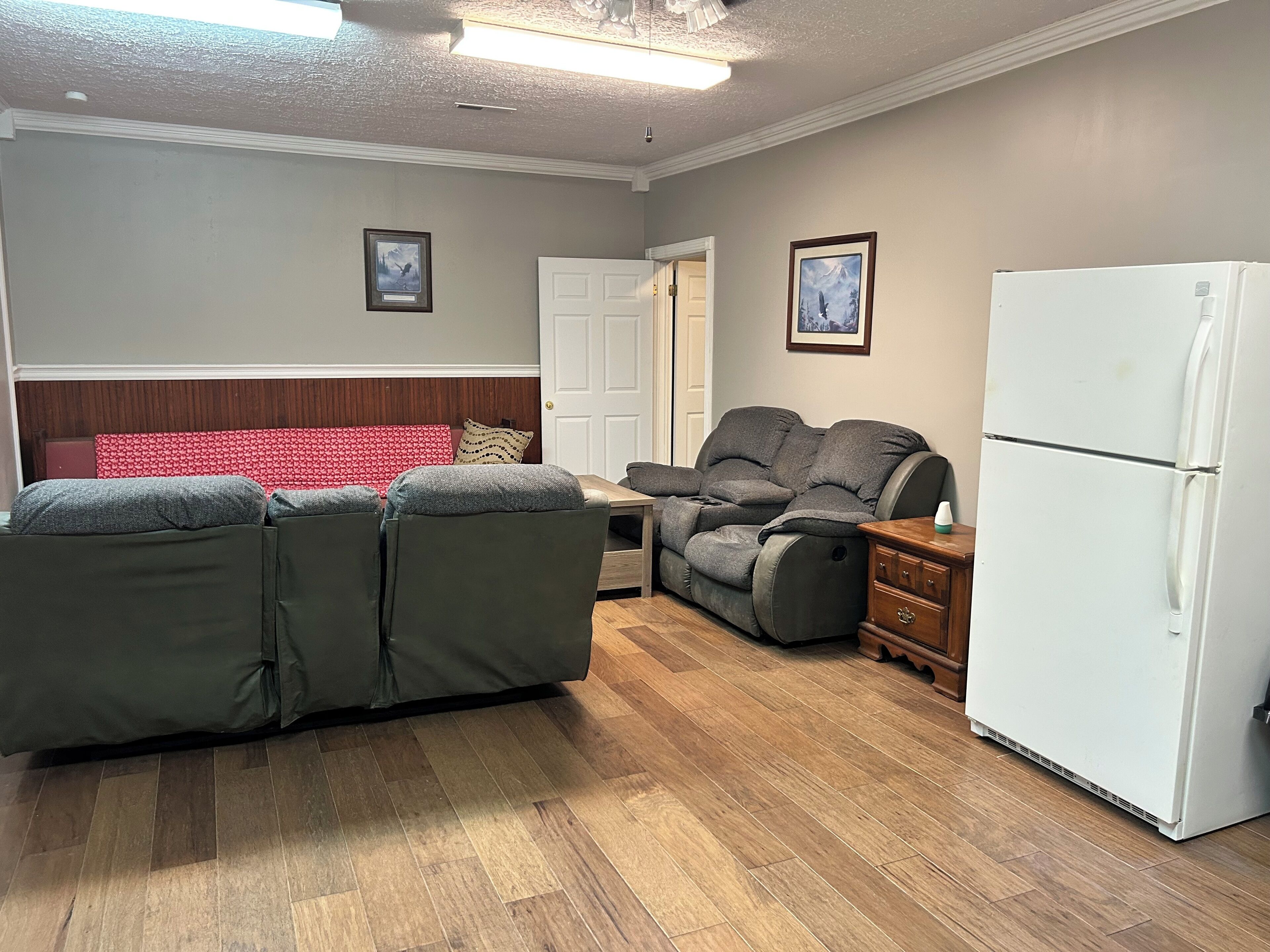 Un salon comprenant un canapé d’angle, un fauteuil inclinable, un réfrigérateur blanc et un plancher en bois.